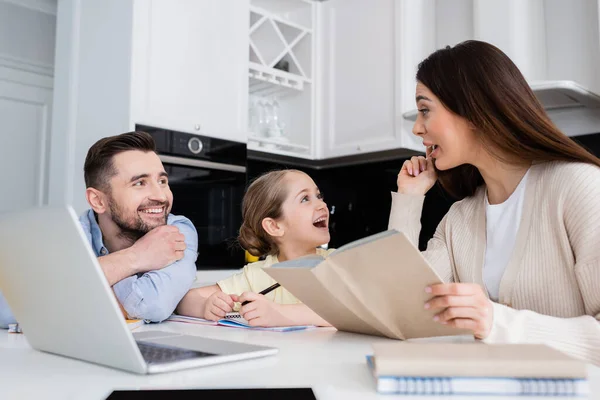 Женщина показывает жест идеи, помогая дочери делать домашнее задание с улыбающимся мужем — стоковое фото