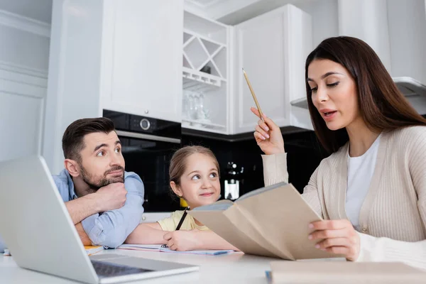 Woman reading book while helping daughter doing homework near husband — Stock Photo
