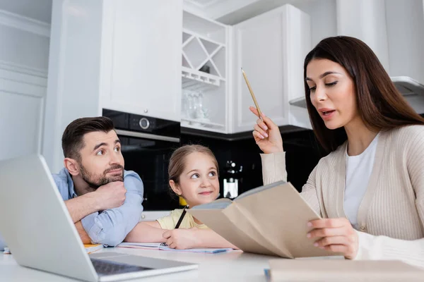 Mujer leyendo libro mientras ayuda a hija haciendo la tarea cerca de marido - foto de stock