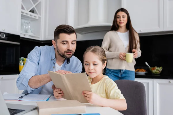 Homme pointant vers le livre tout en faisant des devoirs avec la fille près de la femme souriant sur fond flou — Photo de stock