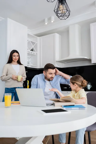 Fille pointant avec la main tout en faisant des devoirs avec père confus près de maman souriante — Photo de stock