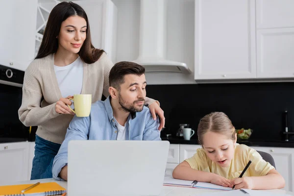 Mädchen schreibt in Notizbuch, während sie Hausaufgaben in der Nähe von Papa und lächelnder Mutter macht — Stockfoto