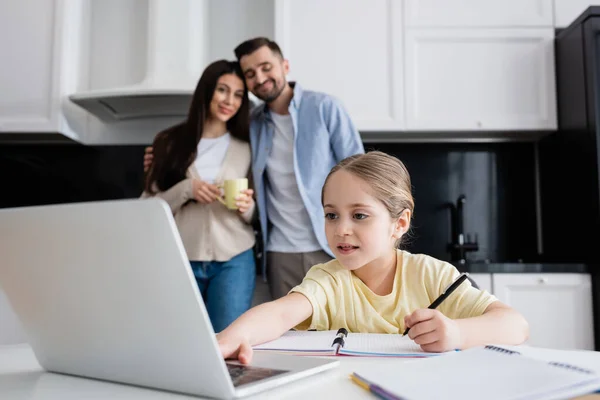 Kid using laptop while doing homework near parents smiling on blurred background — Stock Photo