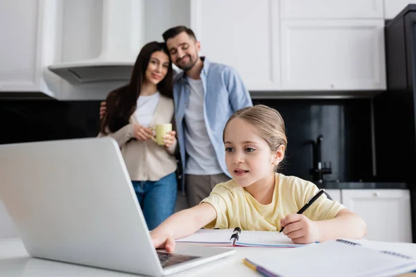 Criança usando laptop ao fazer lição de casa perto dos pais sorrindo no fundo embaçado — Fotografia de Stock