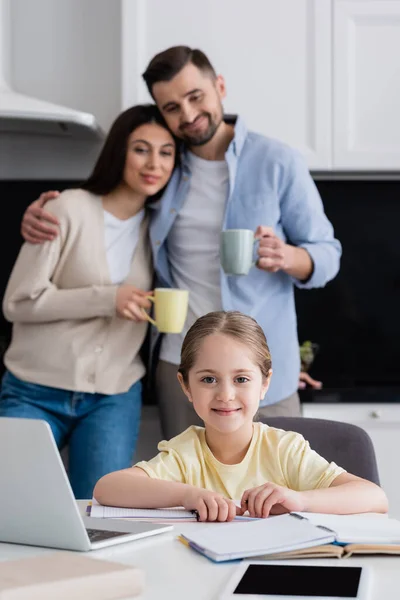Girl smiling at camera while doing homework near happy parents on blurred background — Stock Photo