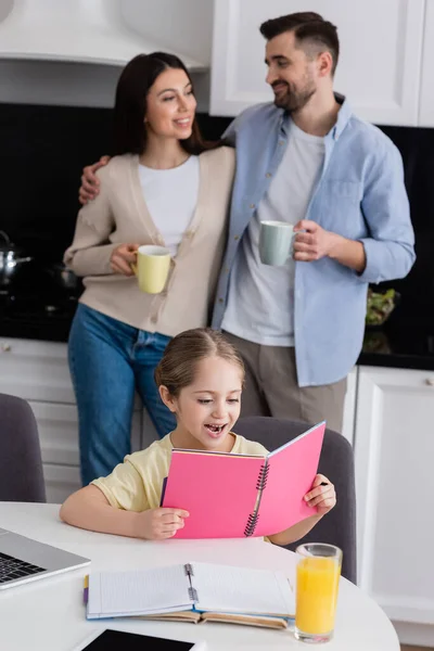 Girl reading notebook while doing homework near happy parents hugging on blurred background — Stock Photo