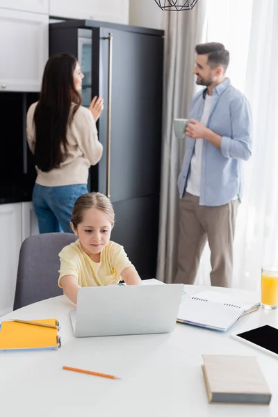 Дівчина друкує на ноутбуці, роблячи домашнє завдання біля батьків, говорячи на розмитому фоні — стокове фото