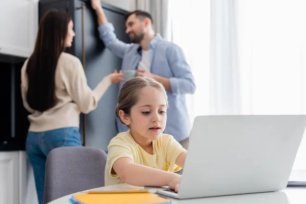 Blurred couple talking near child typing on laptop while doing homework — Stock Photo