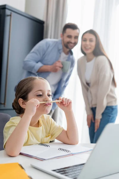 Menina pensativa fazendo lição de casa perto dos pais sorrindo no fundo borrado — Fotografia de Stock