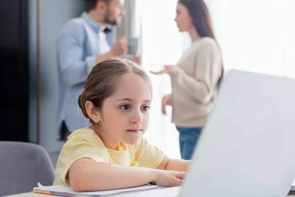 Chica concentrada usando el ordenador portátil mientras hace la tarea cerca de los padres hablando en un fondo borroso - foto de stock