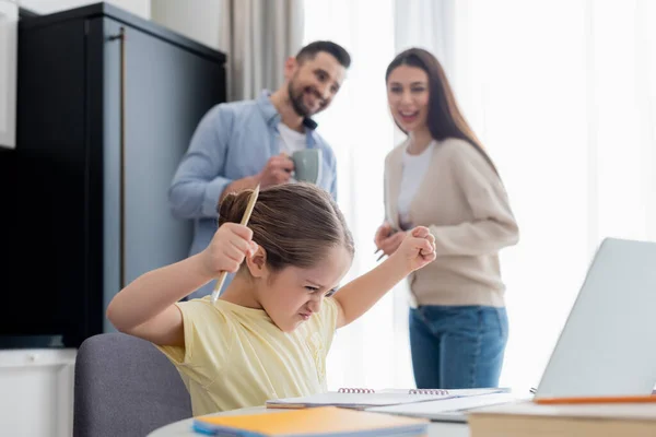 Blurred couple smiling near angry girl showing clenched fists while doing homework — Stock Photo
