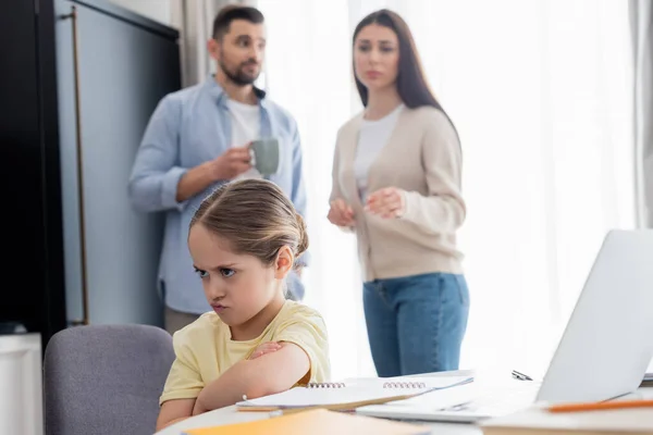 Chica ofendida sentada con brazos cruzados cerca de padres preocupados sobre fondo borroso - foto de stock