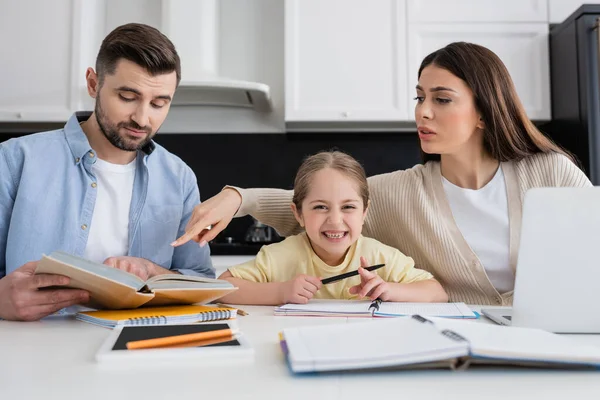 Woman pointing at book in hands of husband while helping cheerful daughter doing homework — Stock Photo