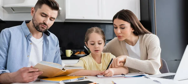 Hombre leyendo libro de texto y mujer señalando con el dedo mientras hace la tarea con la hija, bandera - foto de stock