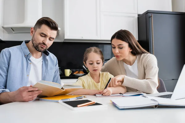 Man reading book and woman pointing with finger while helping daughter doing homework — Stock Photo
