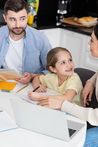 Marido y mujer mirándose el uno al otro mientras ayudan a la alegre hija con la tarea - foto de stock