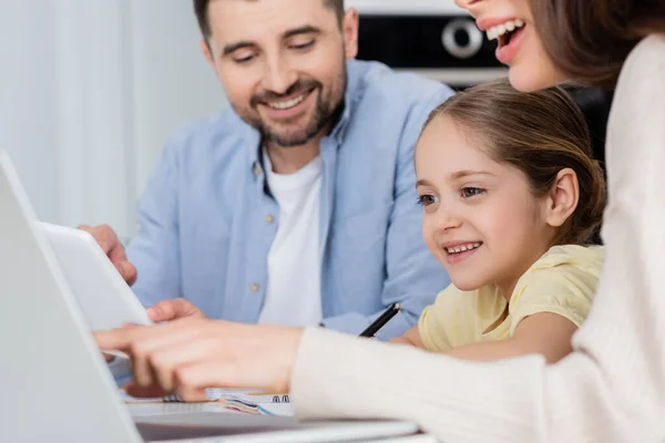 Mujer borrosa apuntando a la computadora portátil cerca de hija y marido sonriente - foto de stock