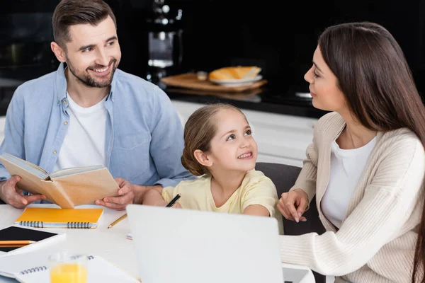 Lächelnde Eltern, die sich bei den Hausaufgaben mit ihrer fröhlichen Tochter anschauen — Stockfoto