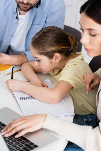 Chica escribiendo en el cuaderno cerca de papá y la madre borrosa utilizando el ordenador portátil - foto de stock