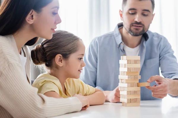 Blurred man removing block from wooden tower near wife and daughter — Stock Photo