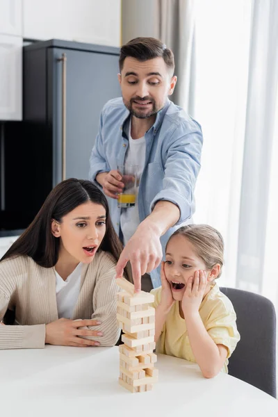 Uomo con succo d'arancia mettendo blocco su torre di legno vicino a moglie e figlia stupite — Foto stock