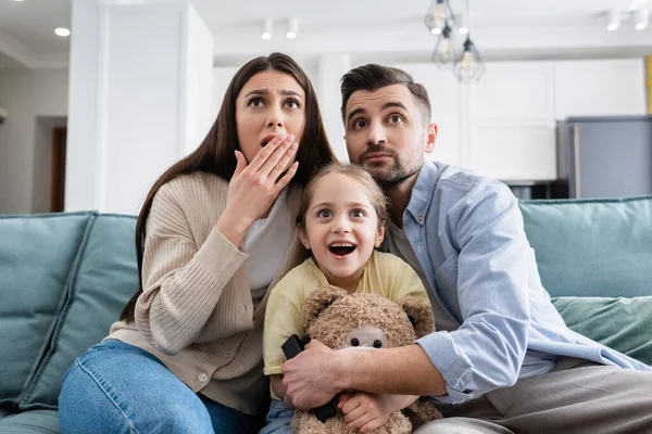 Erstauntes Mädchen schaut Film in der Nähe schockierter Eltern und Teddybär — Stockfoto