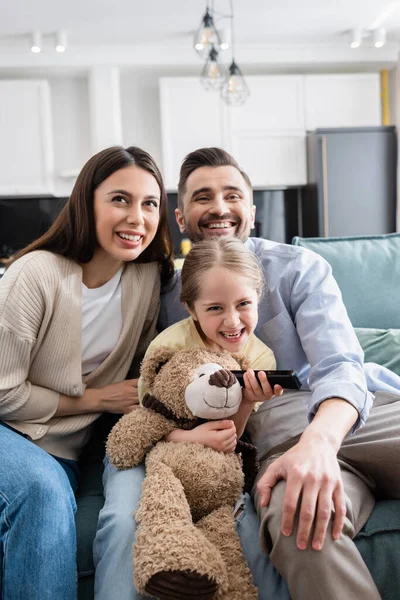 Alegre chica sosteniendo mando a distancia mientras mira la televisión con feliz padres - foto de stock