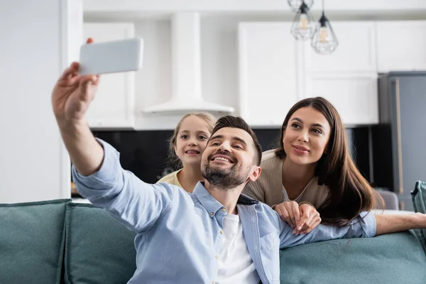 Веселый мужчина делает селфи на мобильном телефоне с улыбающейся семьей дома — стоковое фото