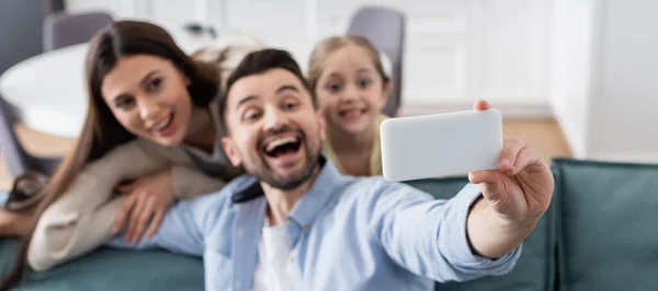 Foco seletivo de smartphone na mão de homem alegre que toma selfie com esposa feliz e filha, bandeira — Fotografia de Stock