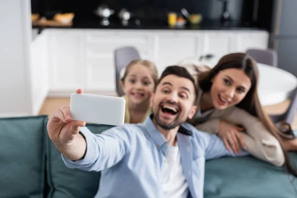 Enfoque selectivo del teléfono celular en la mano del hombre excitado tomando selfie con la familia feliz - foto de stock