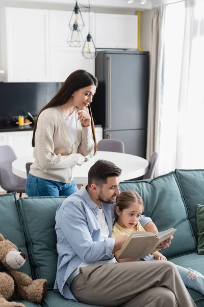 Woman standing near husband and daughter reading book on sofa at home — Stock Photo