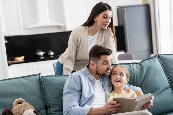 Frau schaut glückliche Tochter und Ehemann beim Lesen von Buch auf Couch an — Stockfoto