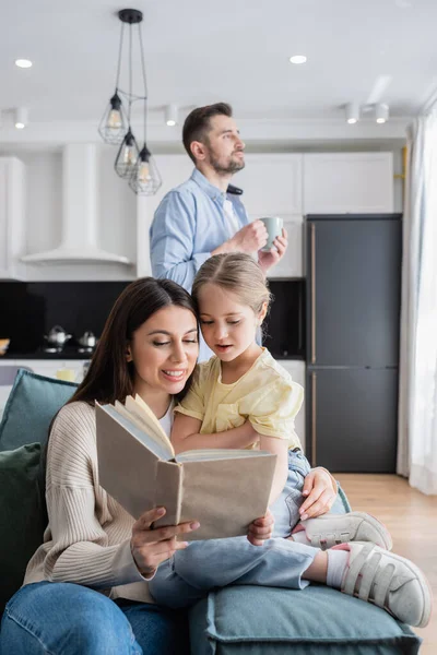 Femme souriante lecture livre à fille près de mari avec tasse de thé — Photo de stock