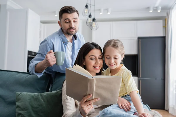 Homem com xícara de chá olhando para esposa e filha lendo livro juntos — Fotografia de Stock