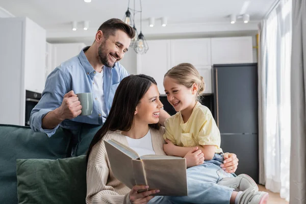 Alegre madre e hija mirándose mientras lee libro cerca de hombre feliz con taza de té - foto de stock