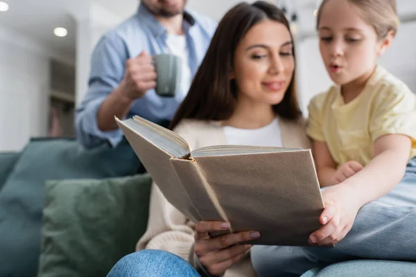 Selective focus of book in hand of woman reading to daughter near blurred husband — Stock Photo
