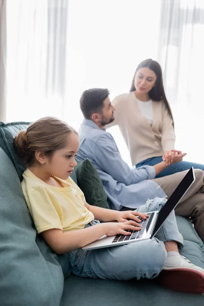 Fille tapant sur ordinateur portable près des parents parlant et tenant la main sur fond flou — Photo de stock