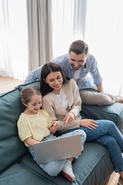 Щасливий чоловік біля веселої дружини і дочки вказує на ноутбук, сидячи на дивані — стокове фото