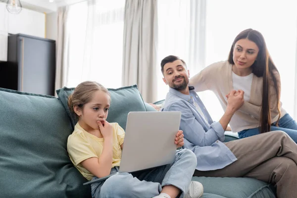 Smiling parents looking at thoughtful daughter using laptop on sofa at home — Stock Photo