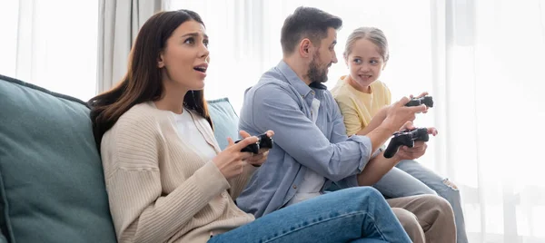 KYIV, UCRANIA - 8 de abril de 2021: mujer tensa jugando con joystick cerca de la familia, pancarta - foto de stock