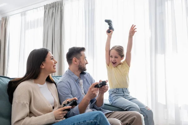 QUIIV, UCRÂNIA - 8 de abril de 2021: menina alegre com joystick mostrando gesto de vitória perto de pais felizes em casa — Fotografia de Stock