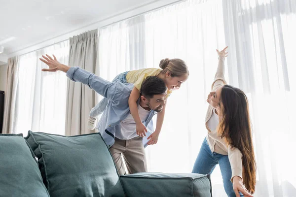 Alegre chica piggybacking en padres espalda cerca de madre imitando avión - foto de stock