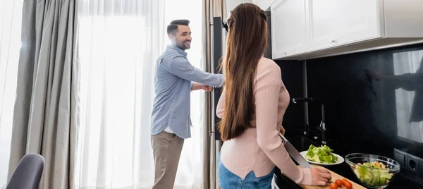 Smiling man opening fridge near wife preparing breakfast in kitchen, banner — Stock Photo