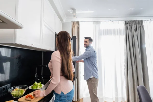 Homem feliz que abre o refrigerador perto da esposa que corta tomates de cereja para a salada — Fotografia de Stock