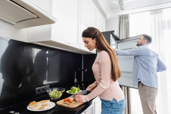 Uomo apertura frigorifero vicino moglie taglio pomodorini vicino filetto di pollo alla griglia — Foto stock