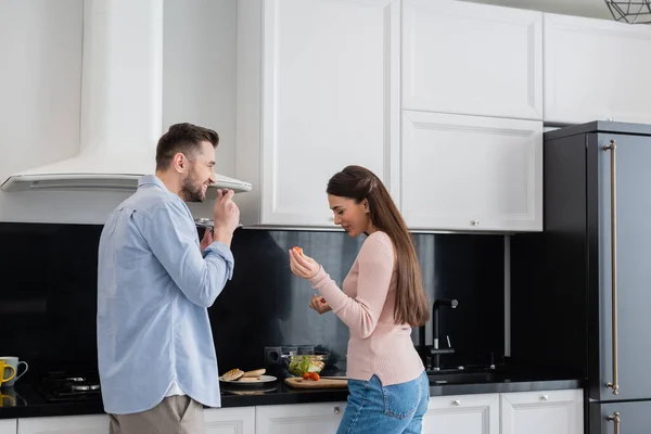 Homem alegre segurando tampa panela perto de esposa feliz preparando café da manhã — Fotografia de Stock