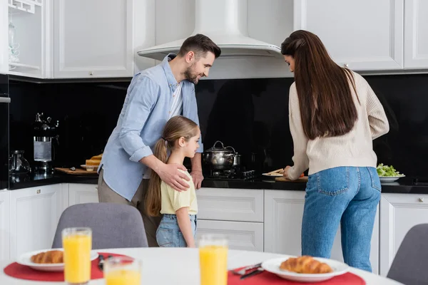 Hombre con hija mirando a la esposa preparando el desayuno cerca de jugo de naranja y croissants en primer plano borrosa - foto de stock