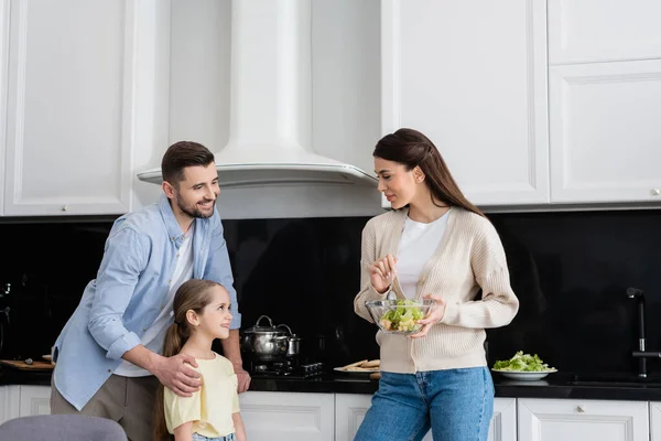 Uomo sorridente abbracciando figlia vicino moglie mescolando insalata di verdure in cucina — Foto stock