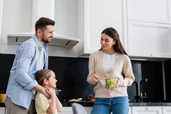 Donna sorridente che tiene la ciotola d'insalata vegetale vicino a marito e moglie in cucina — Foto stock