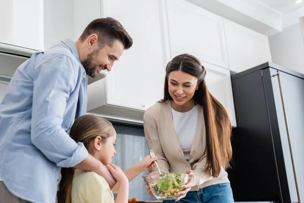 Glücklicher Mann umarmt Tochter beim Mischen von Gemüsesalat neben lächelnder Mutter — Stockfoto